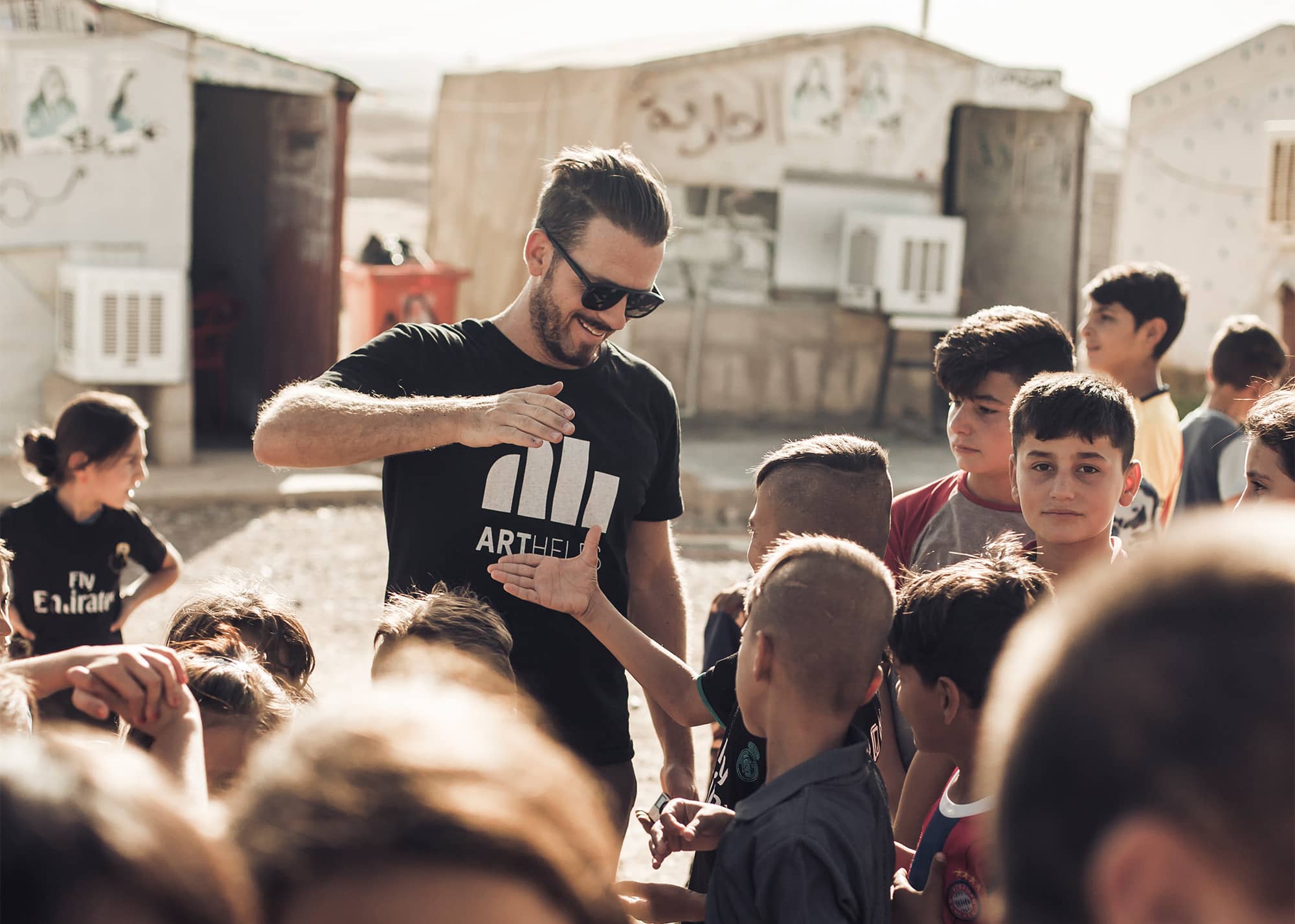 In a refugee camp in Iraq, a young ARTHELPS staff member stands in front of three temporary shelters, greeting a group of smiling, expectant children gathered around him.