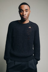 1c1y ss23 one macro summer knit in black distressed look upper detail of male model skin is showing through the ripped part on upper chest left both hands in pockets showing the relaxed look of the sweater slightly stacking on chest and sleeves